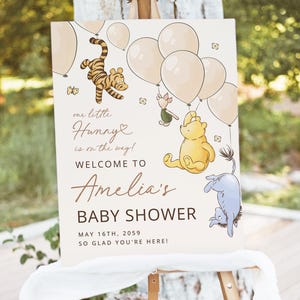 Pode incluir: Uma placa de ch&aacute; de beb&ecirc; com personagens de Winnie the Pooh e bal&otilde;es. A placa diz "our little Hunny is on the way! Welcome to Amelia's Baby Shower May 16th, 2059 So glad you're here!" em um fundo branco.