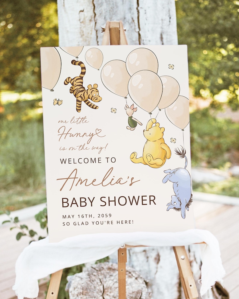 Pode incluir: Uma placa de ch&aacute; de beb&ecirc; com personagens de Winnie the Pooh e bal&otilde;es. A placa diz "our little Hunny is on the way! Welcome to Amelia's Baby Shower May 16th, 2059 So glad you're here!" em um fundo branco.