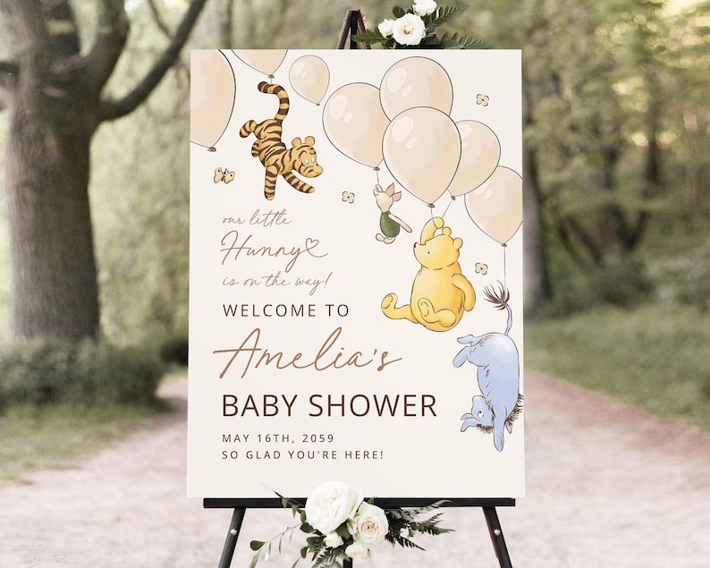 Pode incluir: Placa de boas-vindas para ch&aacute; de beb&ecirc; com personagens do Ursinho Pooh, bal&otilde;es e o texto "Our little Hunny is on the way!" e "Welcome to Amelia's Baby Shower". A placa est&aacute; em um cavalete.
