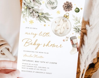 May include: A baby shower invitation with a watercolour design. The invitation features the text "merry little Baby shower" in gold script, along with decorative elements like baubles, greenery, and a baby onesie. The invitation is held by a hand.