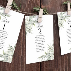 May include: White table cards with elegant floral designs and guest names, suspended by clothespins on a string. The cards are labeled with table numbers and feature names like Chloe Lopez and Madison Wilson. The background is a dark wood.