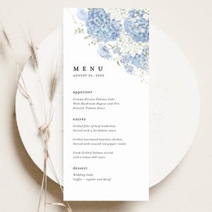 May include: A white menu card with blue and white floral accents. The menu includes appetizers, entrées, and dessert options. The date August 25, 2059, is displayed at the top. The menu is on a white plate with dried grass.