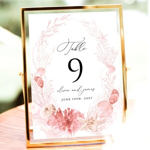 May include: A gold-framed table number card with a watercolor design. The card features the number "9" in black, surrounded by a wreath of coral, seashells, and starfish in shades of pink. The names "Olivia and James" and the date "June 19th, 2057" are also displayed.