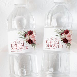 Peut inclure: Deux bouteilles d'eau en plastique transparent avec des étiquettes indiquant "Amelia's Bridal Shower" avec un motif floral dans les tons bordeaux, rose et vert. Le texte sur les étiquettes indique également "August 19, 2026".