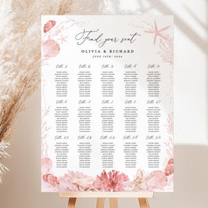 Puede incluir: Un plano de asientos de boda en un caballete. El letrero blanco presenta las palabras "Find your seat" y los nombres Olivia & Richard, con la fecha 19 de junio de 2056. El diseño incluye conchas marinas rosas y acentos de coral.