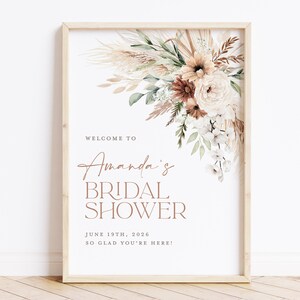 Op de afbeelding: Een ingelijste bruidsdouche-bord met een bloemstuk rechtsboven. Het bord leest "Welkom bij Amanda's Bridal Shower" met de datum 19 juni 2026 en de zin "Zo blij dat je er bent!"