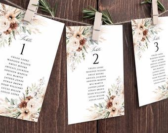 May include: Three white table cards with floral arrangements and guest names, hanging from a string with clothespins. The cards feature elegant calligraphy and a rustic, bohemian design with pampas grass and neutral tones.