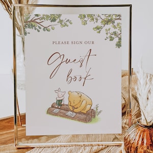 May include: A white sign with green leaves at the top and a gold frame. The sign says "Please sign our Guest Book" in brown text. There is a drawing of Winnie the Pooh and Piglet writing in a log book.