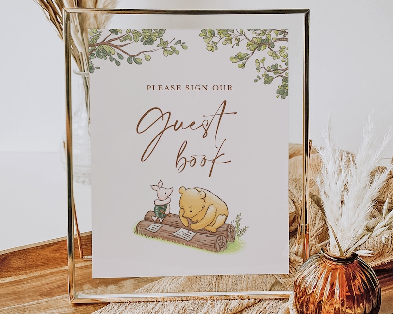 May include: A white sign with green leaves at the top and a gold frame. The sign says "Please sign our Guest Book" in brown text. There is a drawing of Winnie the Pooh and Piglet writing in a log book.