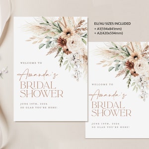 Op de afbeelding: Twee witte rechthoekige borden met bloemstukken en tekst. De borden luiden "Welkom bij Amanda's Bridal Shower" met de datum "19 juni 2026". Het grotere bord bevat EU/AU-maatinformatie. De bloemstukken bevatten beige, bruine en witte bloemen.