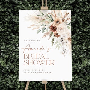 Op de afbeelding: Een wit bord op een houten ezel met bloemstukken en de tekst "Welkom bij Amanda's Bridal Shower, 19 juni 2026, Zo blij dat je er bent!" Het bloemstuk bevat pampasgras, rozen en groen.