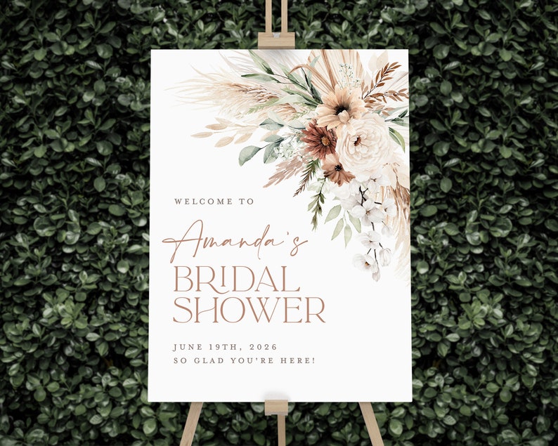 Op de afbeelding: Een wit bord op een houten ezel met bloemstukken en de tekst "Welkom bij Amanda's Bridal Shower, 19 juni 2026, Zo blij dat je er bent!" Het bloemstuk bevat pampasgras, rozen en groen.