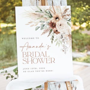 Op de afbeelding: Een wit bord op een ezel, versierd met bloemstukken in neutrale tinten. Het bord leest "Welcome to Amanda's Bridal Shower", de datum 19 juni 2026 en de zin "So glad you're here!"