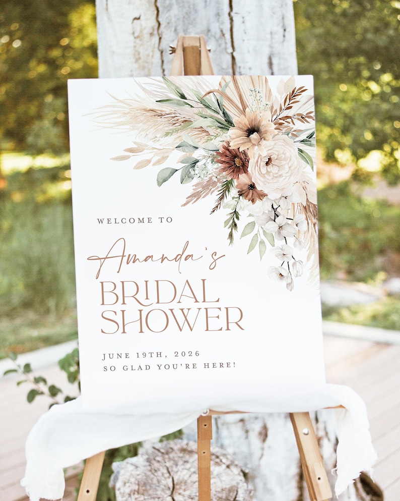 Op de afbeelding: Een wit bord op een ezel, versierd met bloemstukken in neutrale tinten. Het bord leest "Welcome to Amanda's Bridal Shower", de datum 19 juni 2026 en de zin "So glad you're here!"