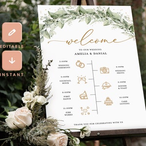 Op de afbeelding: Een wit welkomsbord voor een bruiloft met groen blad en gouden accenten. Het bord vermeldt de huwelijksceremonie, de cocktailtijd, de eerste dans, het aansnijden van de taart en het vuurwerk. De tekst luidt "Welkom op onze bruiloft Amelia & Danial".