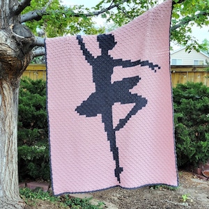 May include: A rectangular crochet blanket featuring a pixelated black silhouette of a ballet dancer in a pose against a solid light pink background. The blanket has a dark gray border and is constructed with a textured, repeating stitch pattern.
