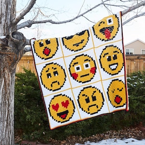 Puede incluir: Una manta blanca de crochet con una cuadrícula de caras de emoji amarillas. Los emojis incluyen una cara sonriente con corazones por ojos, una cara fruncida, una cara sorprendida y una cara guiñando un ojo.