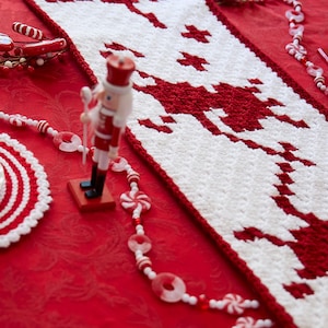 May include: White and red crocheted table runner with a Christmas tree design. The runner is on a red tablecloth with a nutcracker figurine and candy cane decorations.