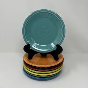 May include: A stack of colorful ceramic plates in various shades, including teal, orange, red, yellow, and blue. The plates are displayed on a black stand, showcasing their vibrant colors and classic design. The plates are a standard size for everyday use.