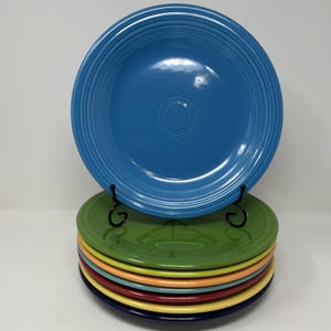 May include: A stack of colorful ceramic plates in various shades, including blue, green, yellow, and red. A blue plate is displayed on a black metal stand. The plates have a classic, ridged design.