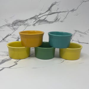 May include: Five colorful ceramic bowls in shades of yellow, green, teal, and gold are stacked. The bowls have a rounded shape with a slightly flared rim. The bowls are arranged on a white marble-patterned surface.