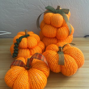 May include: Four orange knit pumpkins with textured fabric surfaces. Each pumpkin features a brown cinnamon stick stem. One pumpkin is stacked, while the others are individual. Decorative ribbons in green and plaid patterns adorn the stems.