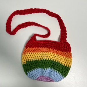 May include: A handmade crochet bag in a rainbow pattern with a red strap. The bag features horizontal stripes of red, orange, yellow, green, blue, and purple. The strap is a solid red color and is approximately 30 inches long.
