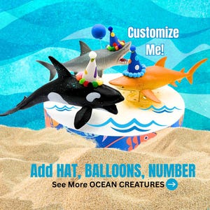 May include: A whimsical cake featuring shark figurines, including an orca, gray shark, and orange shark, each adorned with party hats. The cake is decorated with ocean-themed elements and the text "Customize Me!" and "Add HAT, BALLOONS, NUMBER".