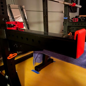 May include: A black metal weightlifting rack with red safety bars and an orange safety clip. A blue towel is draped over the rack.