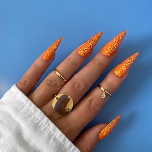 Op de afbeelding: Lange, puntige nagels met oranje glitter en gouden ringen op een hand.