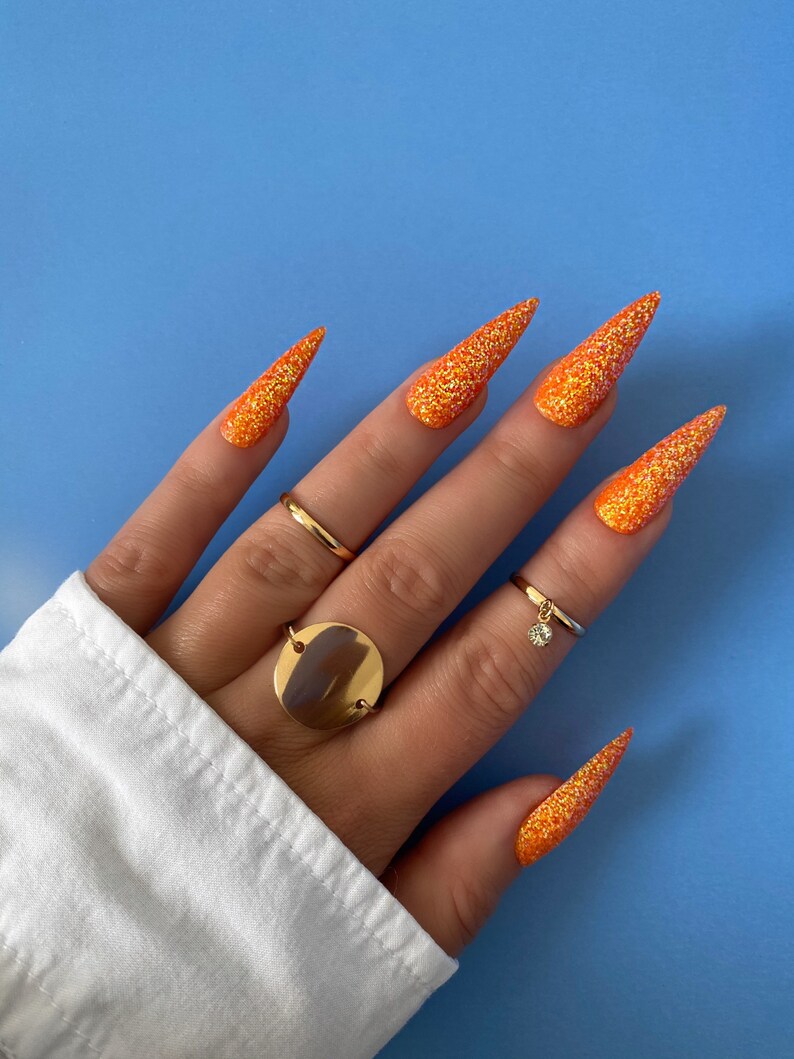 Op de afbeelding: Lange, puntige nagels met oranje glitter en gouden ringen op een hand.