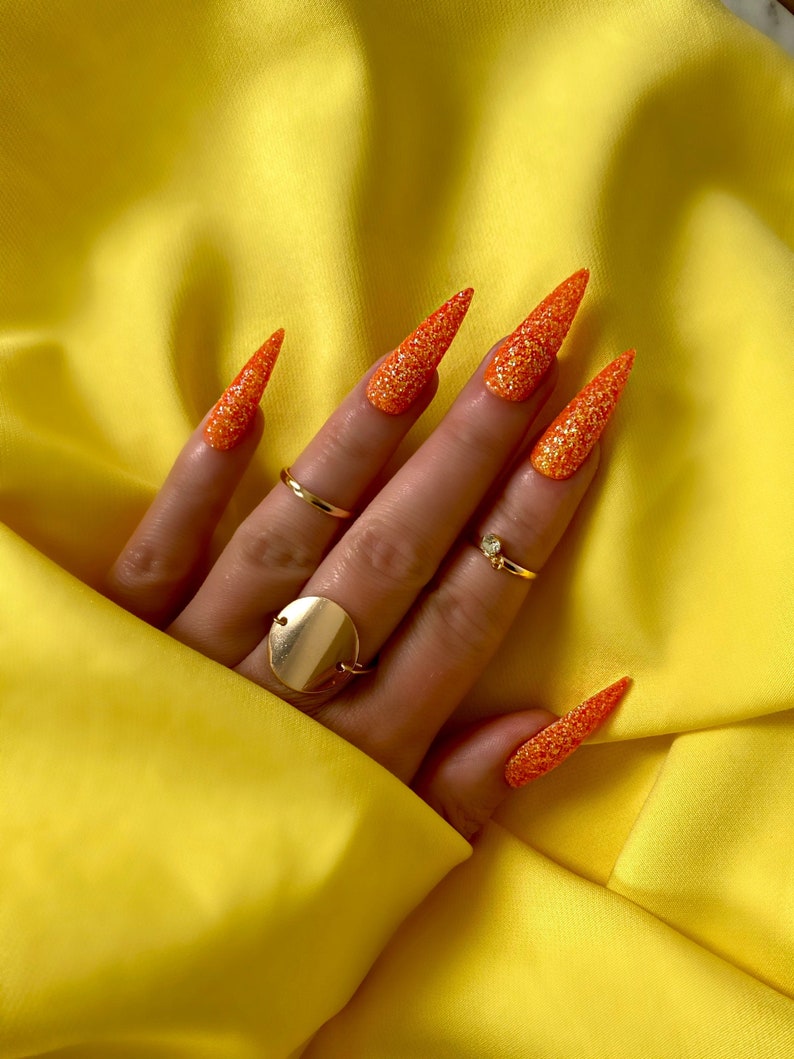 Op de afbeelding: Close-up van een hand met lange, oranje glitternagellak op elke vinger. De nagels zijn in een scherpe punt gevormd. De hand draagt ​​meerdere gouden ringen.