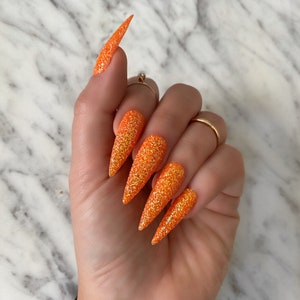 Op de afbeelding: Oranje glitter stiletto valse nagels. De nagels zijn lang en puntig, met een glinsterende afwerking.
