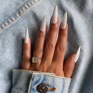 K&ouml;nnte beinhalten: Eine Hand mit langen, wei&szlig;en, glitzernden Stiletto-N&auml;geln. Die Hand tr&auml;gt einen Ring mit einem gro&szlig;en, quadratischen, gelben Edelstein.