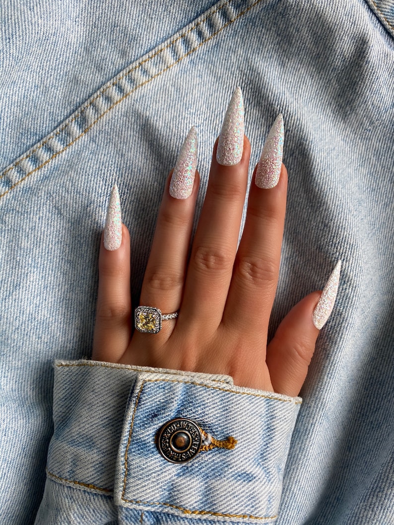 K&ouml;nnte beinhalten: Eine Hand mit langen, wei&szlig;en, glitzernden Stiletto-N&auml;geln. Die Hand tr&auml;gt einen Ring mit einem gro&szlig;en, quadratischen, gelben Edelstein.