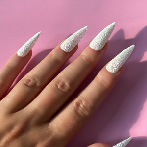 K&ouml;nnte beinhalten: Ein Handmodell mit wei&szlig;en Glitzer-Stiletto-N&auml;geln mit Perlmutt-Finish.