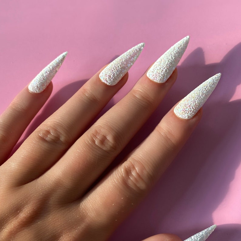 K&ouml;nnte beinhalten: Ein Handmodell mit wei&szlig;en Glitzer-Stiletto-N&auml;geln mit Perlmutt-Finish.