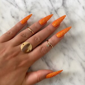 Op de afbeelding: Lange, oranje glitter stiletto nagels met een gouden ring aan de ringvinger.