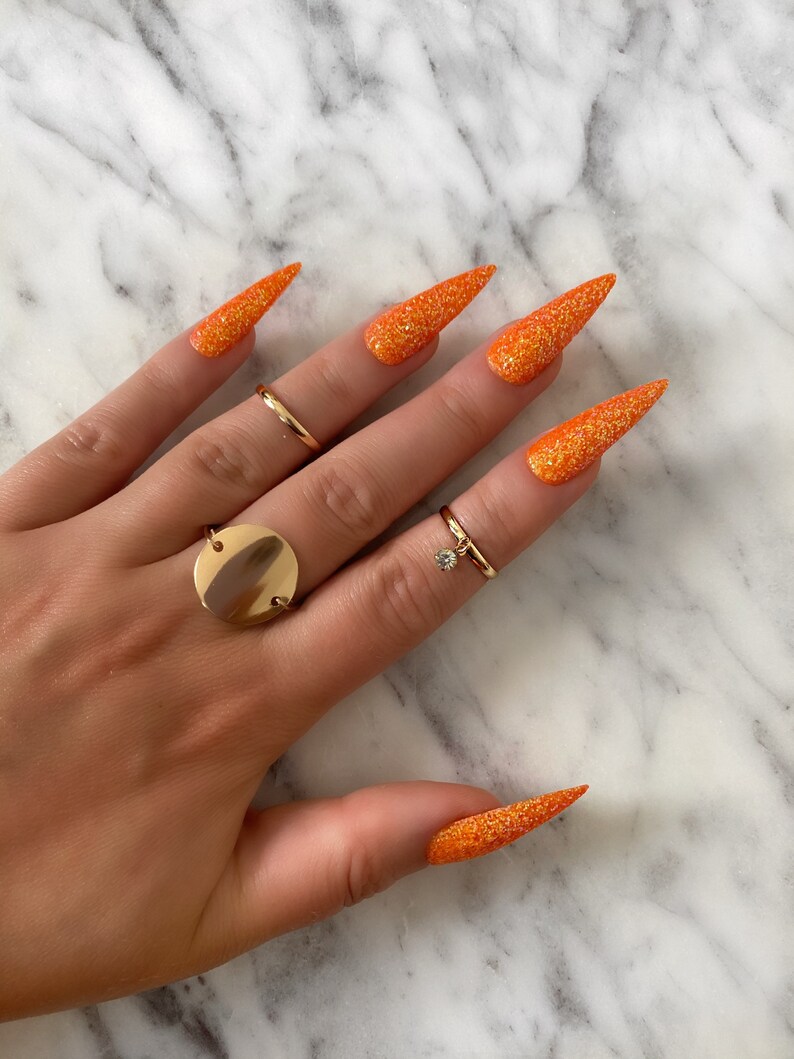 Op de afbeelding: Lange, oranje glitter stiletto nagels met een gouden ring aan de ringvinger.