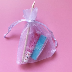 Op de afbeelding: Een witte organza tas met een trekkoord, met daarin twee kleine buisjes met roze en blauwe nagellak. De tas ligt op een roze achtergrond.
