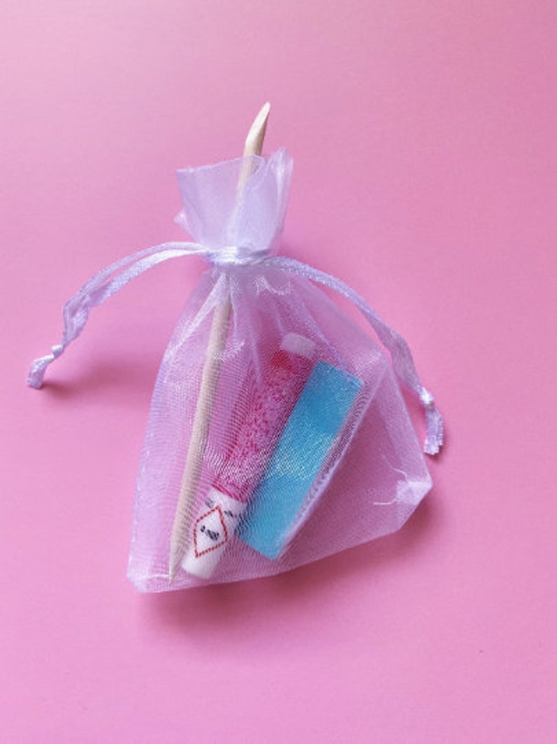 Op de afbeelding: Een witte organza tas met een trekkoord, met daarin twee kleine buisjes met roze en blauwe nagellak. De tas ligt op een roze achtergrond.