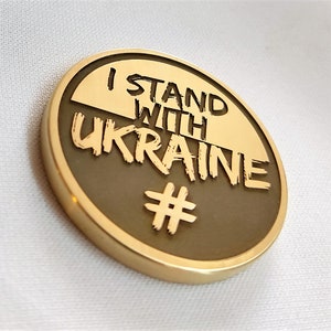 May include: A gold-colored round pin with the words "I STAND WITH UKRAINE" and a hashtag symbol. The pin has a raised design and a polished finish. The text is in a bold, sans-serif font.