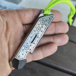 May include: A silver bottle opener with a geometric star pattern and a black tip. It has a slot and a neon green lanyard attached to the top. The bottle opener is held in a hand.