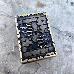 Puede incluir: Un encendedor rectangular de color latón con un diseño detallado. Presenta una calavera emergiendo de una pared de ladrillos, con manos esqueléticas extendidas. El diseño es en tonos negros y grises, con un borde dorado.