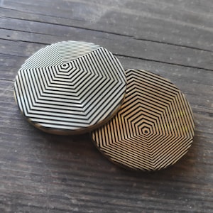 Puede incluir: Dos posavasos de metal de color dorado con un patrón geométrico de hexágonos concéntricos. El patrón es negro y blanco.