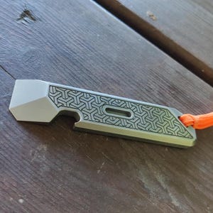 May include: A silver metal multi-tool with a geometric pattern and a bottle opener. The tool has a lanyard loop and is attached to an orange lanyard.