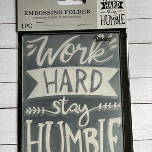 May include: A clear plastic embossing folder with the text "Work Hard Stay Humble" in a white, handwritten font. The folder is packaged in a black and white cardboard sleeve.