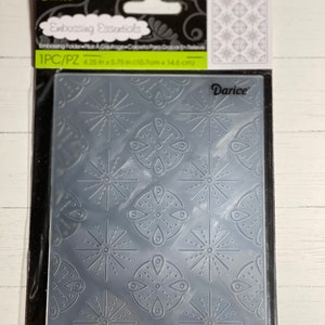 May include: A clear plastic embossing folder with a repeating pattern of stylized snowflakes and circles. The folder is packaged in a clear plastic sleeve with a black header card that reads "Darice Embossing Essentials 1PC/PZ 4.25 in x 5.75 in (10.8 cm x 14.6 cm)"