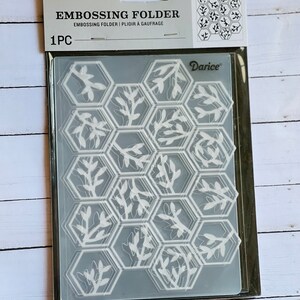 May include: A clear plastic embossing folder with a hexagonal pattern of leaves. The folder is packaged in a clear plastic bag with a white label that reads "Embossing Folder | Plioir à Gaufrage" and "Darice".