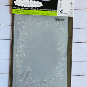 May include: A white plastic embossing folder with a decorative border design. The folder measures 4.25 inches by 5.75 inches (10.7 cm by 14.6 cm).